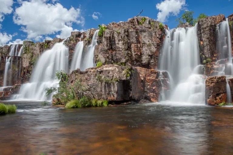 Cachoeira de várias quedas sobre paredão de pedra, com poço de água cor de chá